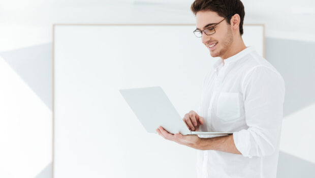 Picture of young man wearing eyeglasses and dressed in white shirt using laptop computer near big board. Look at laptop.