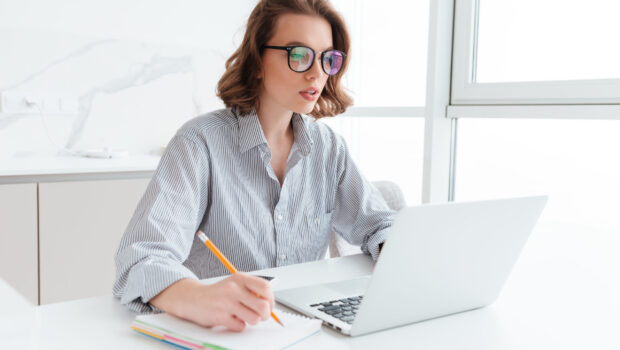 Young concentrated brunette woman in glasses wokking with laptop while sitting at table in light kitchen